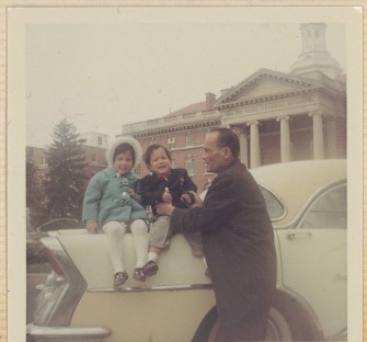 An antique image of two children sitting on a white car with a man holding one of them.